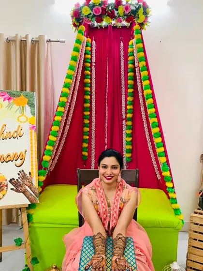 A smiling bride seated in her decorated mehndi ceremony corner, proudly presenting her full bridal henna. We work with you to make your mehndi ceremony picture-perfect.
