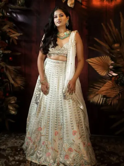 A full-length shot of a model in a white lehenga with subtle floral embroidery, paired with a heavy green necklace.