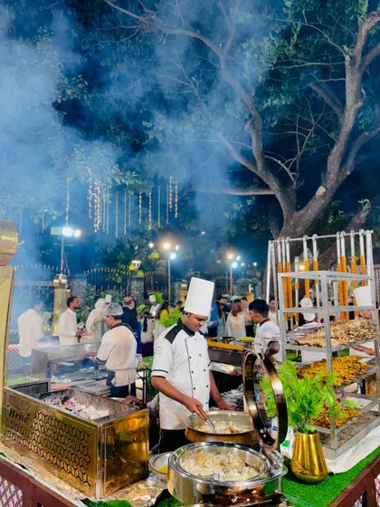 Chefs preparing various grilled items at a smoky barbecue station, with hanging kebabs in the background.
