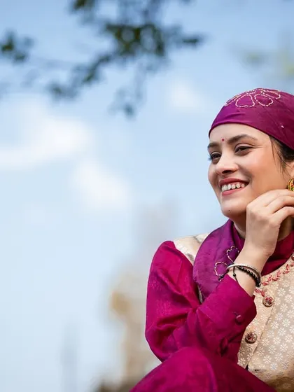 A joyful close-up of the woman adjusting her traditional earring, with a bright blue sky in the background.