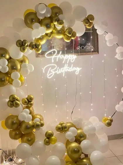 This modern and chic birthday backdrop features a 'Happy Birthday' neon sign inside a circular frame. The frame is asymmetrically decorated with metallic gold and white balloons, with fairy lights adding a warm glow.