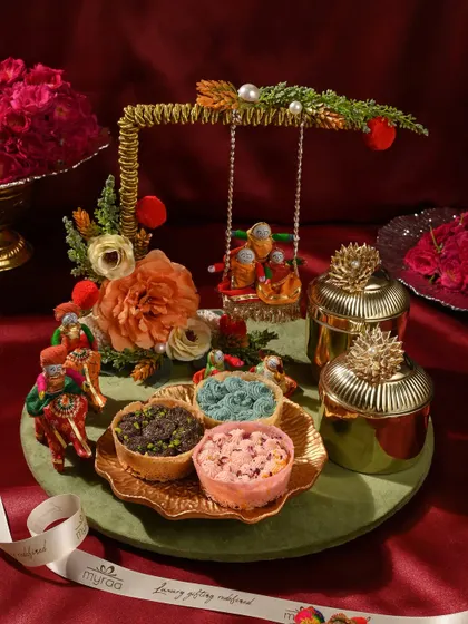 This whimsical Teej hamper features a miniature swing (jhoola), colourful ghewars, and ornate golden jars. It’s a playful and traditional gift that captures the essence of the festival.
