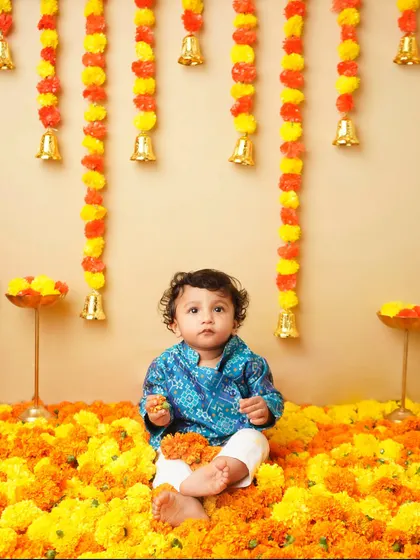A vibrant festive photoshoot of a baby boy dressed in a traditional blue kurta, sitting in a sea of bright orange and yellow marigold flowers, with festive decorations hanging in the background.