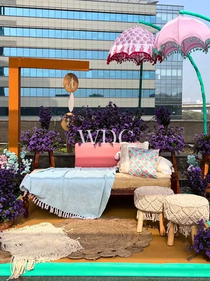 A whimsical and colorful photo-op area. This setup includes a comfortable diwan, macrame stools, and quirky umbrellas, all surrounded by an abundance of purple flowers.