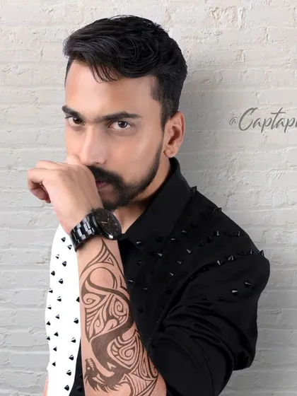 An edgy male portrait with a strong, confident look. The combination of a textured brick background, a split-color shirt, and focused lighting highlights the model's features and the tattoo art, creating a modern fashion photograph.