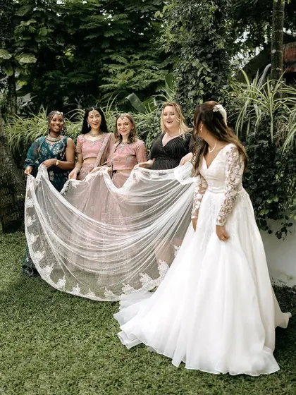 The bride with her international bridesmaids, who are holding her long, beautiful veil. It's a perfect picture of friendship and cultural fusion.
