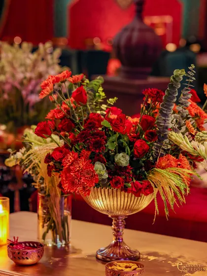 A beautiful floral centerpiece in a fluted golden bowl, surrounded by candlelight. The arrangement of red carnations, roses, and fresh greenery adds a touch of natural elegance to the opulent setting.
