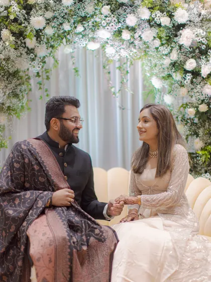 A sweet moment from an engagement ceremony, with the couple seated under a beautiful arch of white flowers, sharing a look of love.