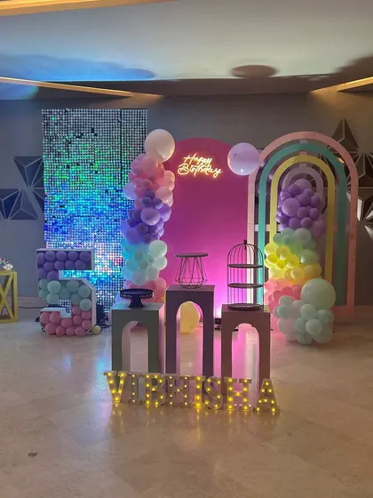 A modern and chic rainbow themed birthday party setup. The decor includes a pink backdrop, a shimmer wall, and artistic rainbow-shaped arches with pastel balloon accents.