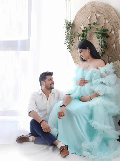 This session features a dreamy, light-filled setup. The expecting mother looks serene in a ruffled, pastel blue gown, sharing a quiet moment with her partner by the window.