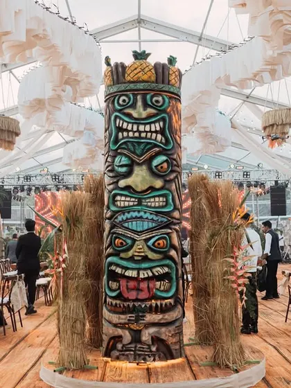 A towering, hand-carved Tiki totem pole stands as a central feature of the event space. Surrounded by pampas grass and rustic decor, it is a powerful and authentic focal point for the tropical theme.