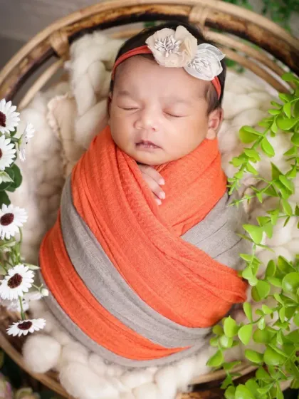 This newborn is peacefully sleeping, swaddled in a lovely orange and grey wrap. We use soft, natural props like this wicker basket and greenery to create a rustic and organic feel.