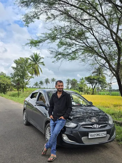 Leaning against my car on a rural road, with fields in the background. My car is my companion on these journeys, taking me to the hidden corners of Karnataka.