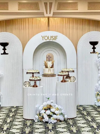 An elegant and serene stage for a First Holy Communion. This setup uses white 3D ridged panels, tall pedestals for the cake and offerings, and lush white and gold floral arrangements for a sophisticated look.