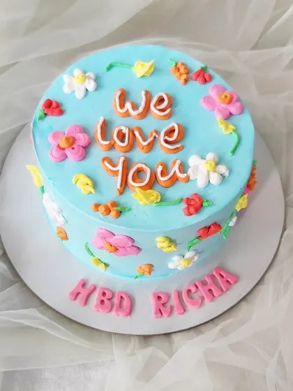 A cheerful and bright minimalist cake with a simple message. The light blue buttercream is decorated with colorful, hand-piped flowers and a "We Love You" message in the center.