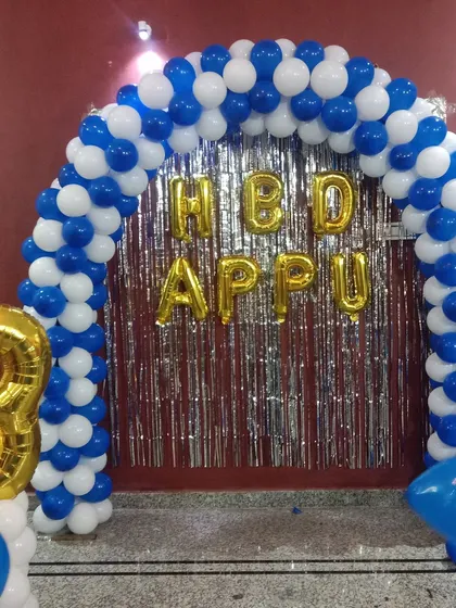 A simple home setup for a third birthday. A blue and white balloon arch frames a silver fringe curtain with 'HBD APPU' in foil letters.