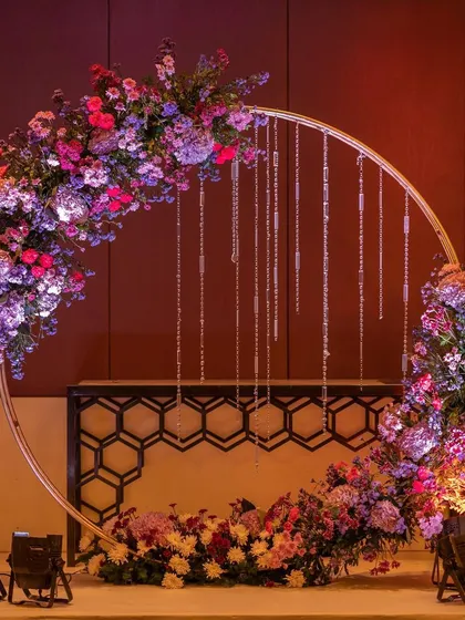 A modern and chic circular photo booth. The golden ring is adorned with a vibrant mix of purple and pink flowers and hanging crystals, creating a stylish spot for guests to capture memories.
