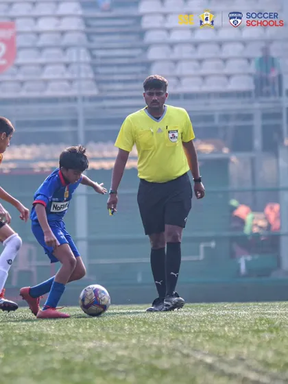 A young player demonstrates composure on the ball under the watchful eye of the referee during an AIFF Youth League match.