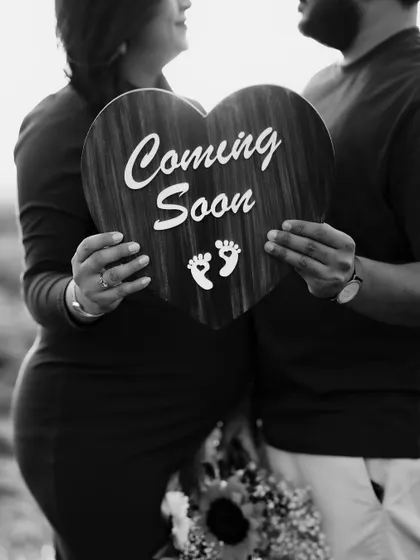 A classic black and white close-up of the couple holding a heart-shaped "Coming Soon" sign. The focus is on their hands and the message, creating a timeless announcement.