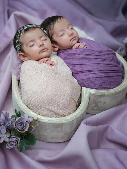 A wider view of the twin session, showing the babies sleeping peacefully in a heart basket surrounded by soft purple fabric and delicate flowers.