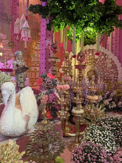 A stunning display of traditional brass lamps and floral arrangements for the Ambani family's Raas Garba. The decor, inspired by Radhe and Krishna, features a sculpted swan and intricate details that evoke a divine atmosphere.