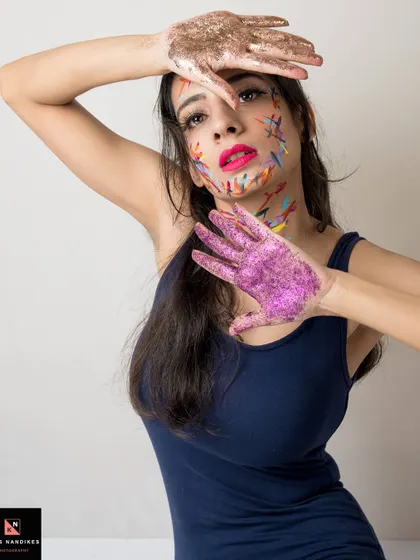 A creative portrait where the model's face and hands are adorned with colorful paint strokes and glitter. Her pose, framing her face, draws attention to the artistic and expressive makeup.