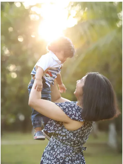 A mother lifts her son towards the sunlit sky in a beautiful park setting. This outdoor family photo captures a moment of pure joy and connection.