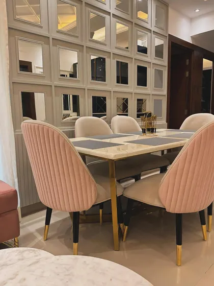 A chic dining area featuring a marble-top table with gold-finished legs. The setup is completed with custom-made pink and beige velvet chairs with channel tufting, set against a stylish wall of mirrors.