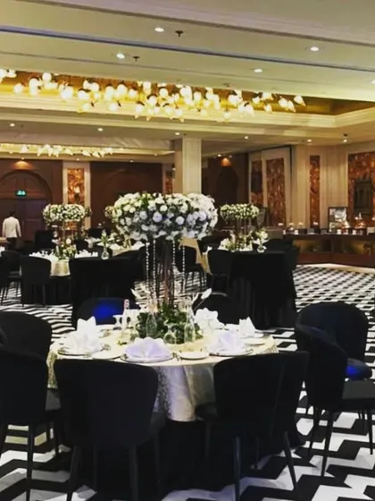 A close-up of a table setting at a modern engagement party. The black velvet chairs and tablecloth are complemented by a tall, elegant centerpiece of white flowers.