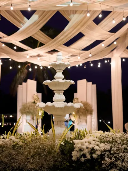 The fountain at night, lit from below and surrounded by glowing flowers under a canopy of bistro lights. The scene is transformed into a magical, romantic focal point.