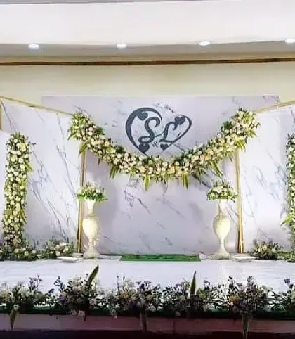 An elegant and clean engagement stage design. The backdrop features white marble-patterned panels with golden frames, decorated with simple yet beautiful arrangements of white flowers and greenery. The couple's monogram adds a lovely personal touch.