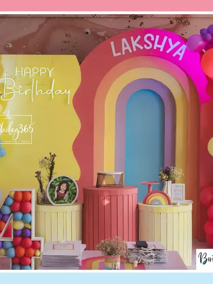 A close-up of the rainbow-themed stage for Lakshya's party, with a "Happy Birthday" neon sign and colorful pedestals.