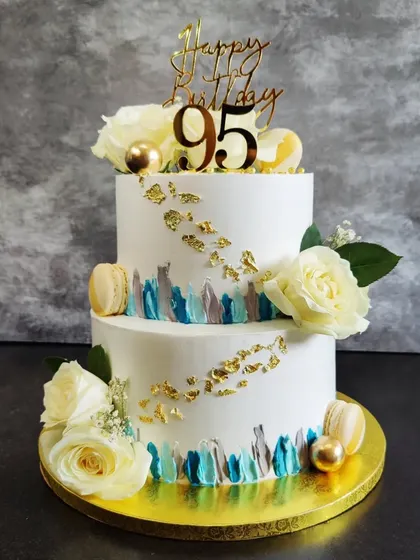 A grand two-tier cake for a 95th birthday. I used a watercolor buttercream technique with blue and grey, accented with gold leaf, white roses, and macarons.