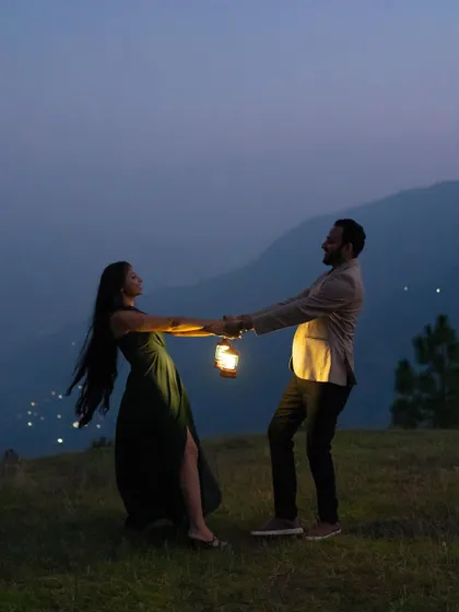 A playful and romantic moment captured during a night shoot in the mountains. The couple dances, holding a lantern whose light catches the movement of her dress, with the distant city lights adding to the magical atmosphere.