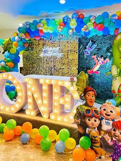 A vibrant first birthday party combining a jungle theme with Cocomelon characters. The stage features large "ONE" marquee letters, a dinosaur cutout, and a colorful balloon arch.