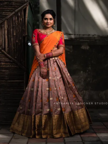 A model wearing a traditional half saree with a brown patterned skirt and a bright orange dupatta. The embroidered red blouse adds a pop of color.