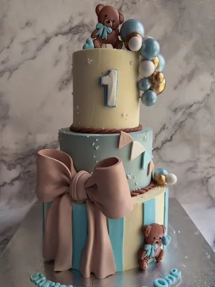 A whimsical two-tier cake perfect for a first birthday. It's adorned with adorable fondant teddy bears, a large sculpted bow, and a balloon garland, creating a storybook feel.