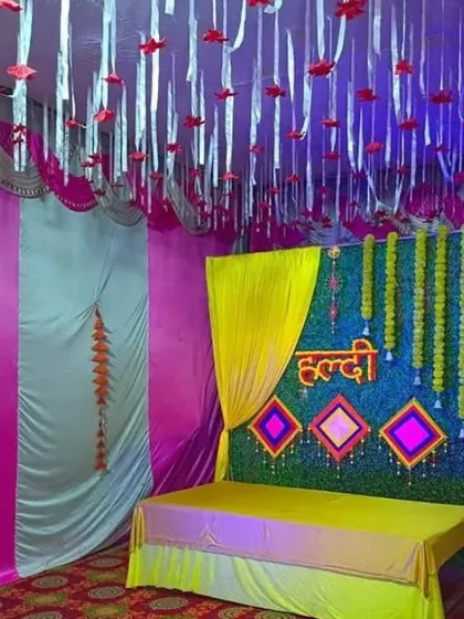 A Haldi stage with a green grass backdrop, yellow draping, and a decorated ceiling with hanging ribbons.