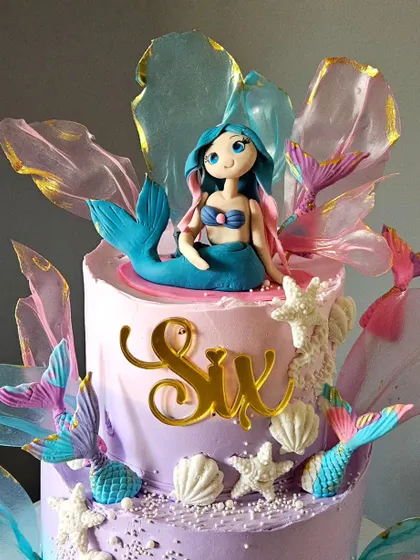 A close-up of the dreamy mermaid cake, highlighting the beautiful, hand-sculpted mermaid topper and the shimmering, edible rice paper sails that mimic fins or waves.