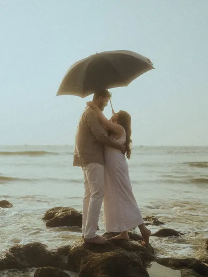 A romantic, movie-like scene of a couple embracing under an umbrella while standing on rocks by the sea. This photo adds a touch of classic romance to their beach photoshoot story.