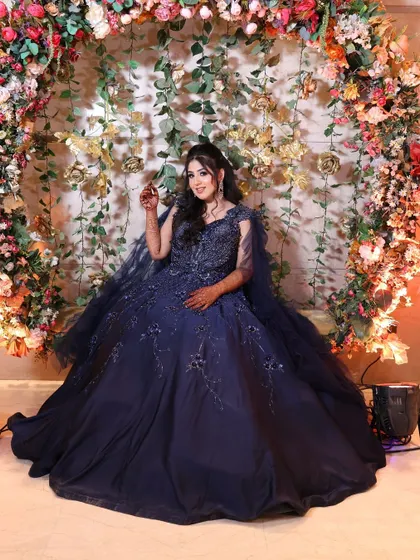 The client posing in front of a floral arch, the gown's full skirt and cape creating a stunning silhouette. A perfect rental for an engagement or reception.