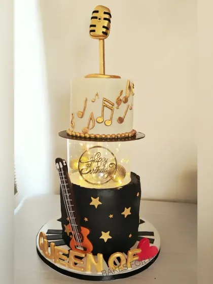 A two-tier music-themed cake for a "Queen of Fitness." The design combines a guitar, piano keys, a microphone, and fairy lights to celebrate her passions.