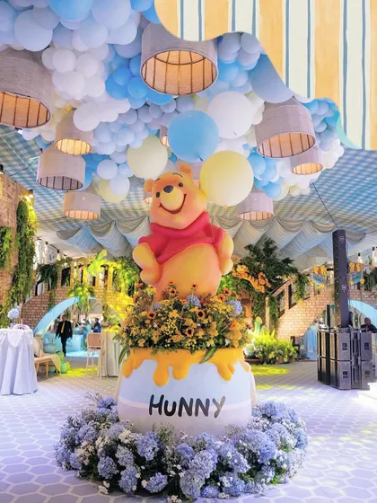 The centerpiece for a Winnie the Pooh first birthday, featuring a larger-than-life Pooh Bear atop a 'Hunny' pot overflowing with sunflowers. The ceiling is a sky of blue and yellow balloons, creating a storybook world.