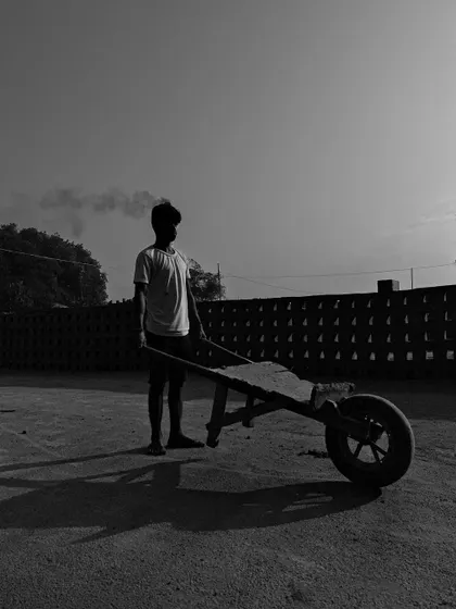 A young boy stands with a wheelbarrow at the brick kiln. This black and white image captures a quiet moment, highlighting the presence of the next generation in these work environments.