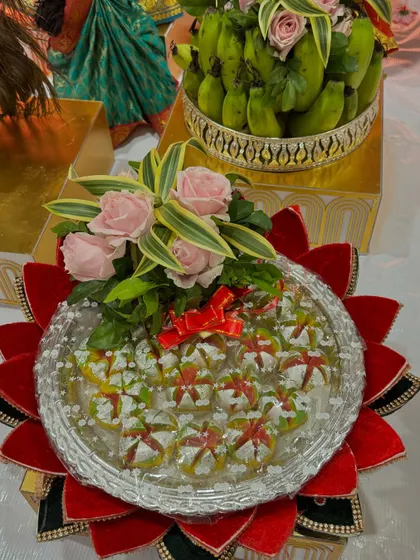 A colorful sweet platter with a red and black lotus base. The sweets are arranged neatly and topped with a small bouquet of pink roses.