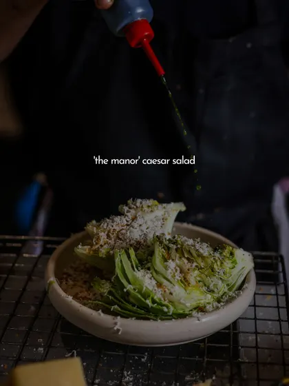 A chef drizzles dressing over the Manor Caesar salad. This action shot highlights the freshness of the ingredients and the care that goes into preparing even the simplest dishes.