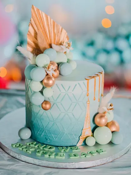 A Paris-themed engagement cake with a geometric pattern on a blue ombre base. The design is adorned with a cascade of blue and gold spheres, a gold Eiffel Tower accent, and dried floral elements.
