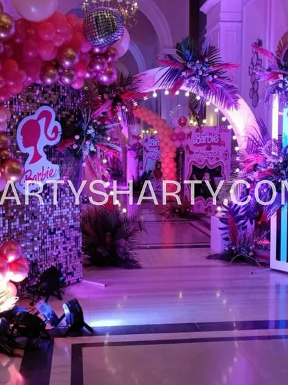 An entrance for a glamorous Barbie-themed party, featuring a sequin wall, tropical leaf arrangements, and a silhouette of the iconic Barbie logo.