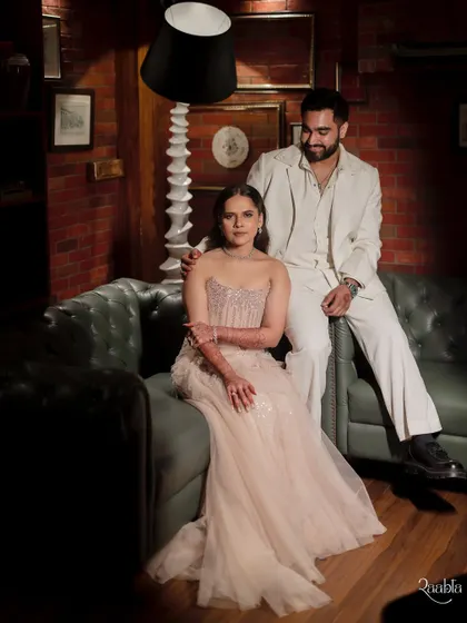 A classic, elegant portrait of influencer Viraj Ghelani and his bride Palak, seated in a moody, sophisticated setting.