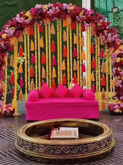 A stunning Mehendi seating arrangement with a vibrant pink sofa set against a backdrop of hanging yellow and orange tassels and a lush floral arch. The traditional carved fire pit adds an authentic touch to the festive decor.